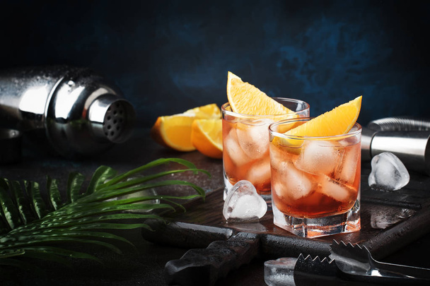 Trendy alcoholic cocktail Negroni with dry gin, red vermouth and red bitter, orange slice and ice cubes. Brown bar counter background, bar tools, night mood, copy space, selective focus - Foto, Imagen