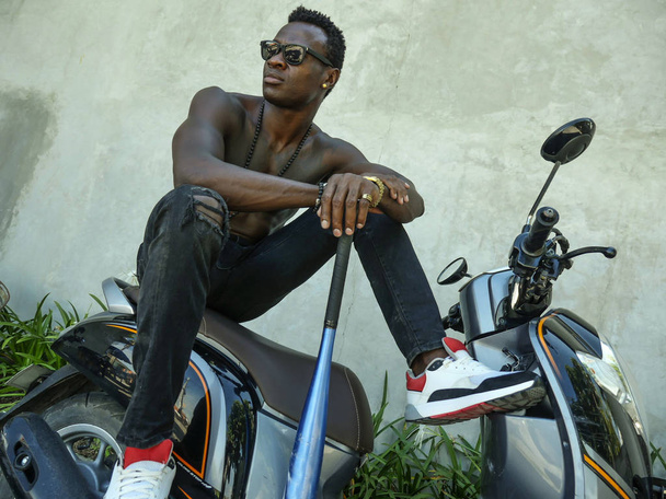 urban lifestyle portrait of fit body and dangerous looking black afro American man with naked torso and sunglasses sitting on scooter holding baseball bat - Foto, immagini