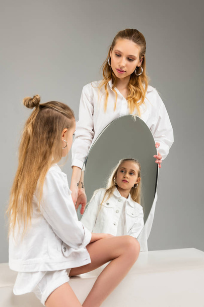 Long-haired little girl in white clothes seriously looking on her reflecting - Foto, Imagem
