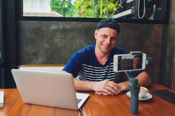 Barbudo atraente blogueiro de vídeo masculino em óculos elegantes atira streaming de vídeo para os usuários enquanto está sentado no café e bebendo café. Masculino vlogger tira fotos de si mesmo na câmera com estabilizador
. - Foto, Imagem