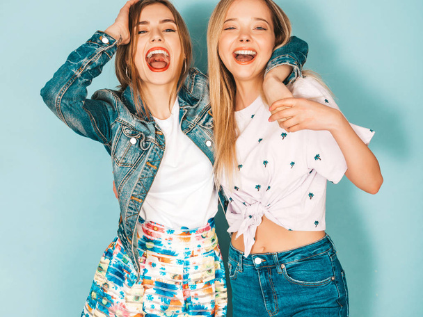 Two young beautiful smiling blond hipster girls in trendy summer colorful  clothes. Sexy carefree women posing near blue wall. Positive models having fun  - Photo, Image