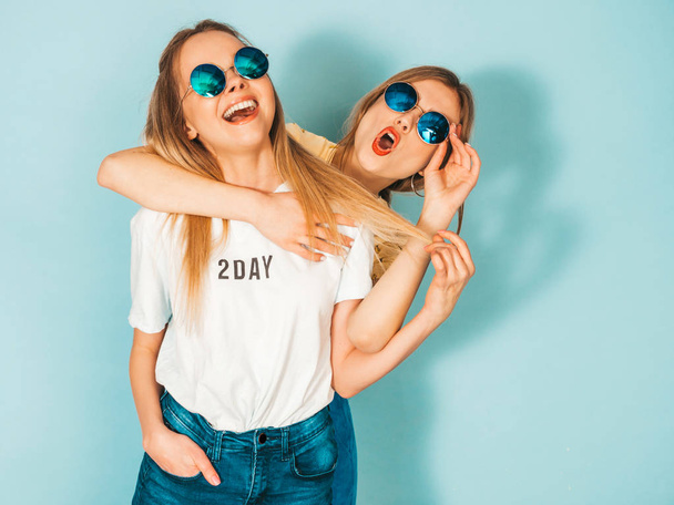 zwei junge schöne lächelnde blonde Hipstermädchen in trendigen sommerfarbenen T-Shirt-Klamotten. sexy unbeschwerte Frauen posieren in der Nähe der blauen Wand mit runden Sonnenbrillen. positive Modelle, die Spaß haben und sich umarmen - Foto, Bild