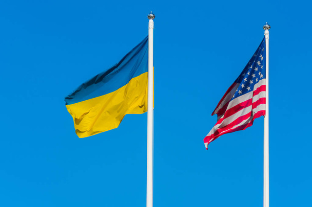 ウクライナの旗とアメリカ合衆国の旗は澄んだ青空の背景に旗竿で互いに横に風に揺れている. - 写真・画像
