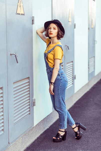 young woman in blue denim overalls and yellow t-shirt with black hat sensual looking at camera while posing near industrial building  - Fotoğraf, Görsel