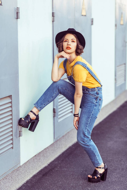 young woman in blue denim overalls and yellow t-shirt with black hat sensual looking at camera while posing near industrial building  - Foto, imagen