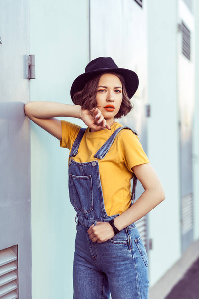 young woman in blue denim overalls and yellow t-shirt with black hat sensual looking at camera while posing near industrial building  - 写真・画像