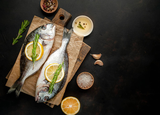 Raw sea bass served with rosemary twigs, lemon slices and spices on wooden board over stone background  - Fotografie, Obrázek