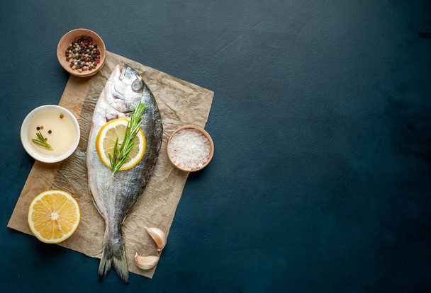 Nyers tengeri sügér szolgált rozmaring gallyak, citromos szelet és a fűszerek a barna papír felett kő háttér  - Fotó, kép