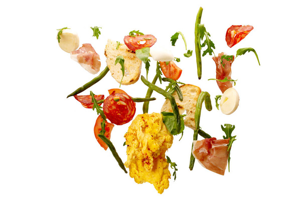 Isolated falling vegetables. Slices of tomato, egg, omelette, salami, prosciutto, asparagus, arugula and cheese. fresh salad ingredients in the air isolated on white background - Photo, Image