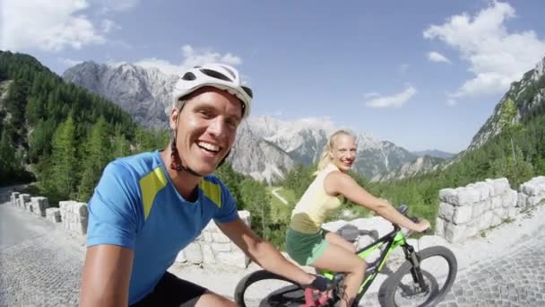 Pomalý pohyb a usmívající se pár na kole na horské cestě za krásným letním dnem. Atraktivní novomanželé, kteří si užijí cyklistiku v horách. Dívka mávala na kameru a šlapává na e-kole. - Záběry, video