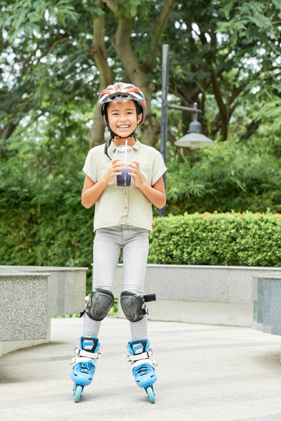 Portrait of happy little roller skater drinking fruit cocktail in summer park - Photo, Image