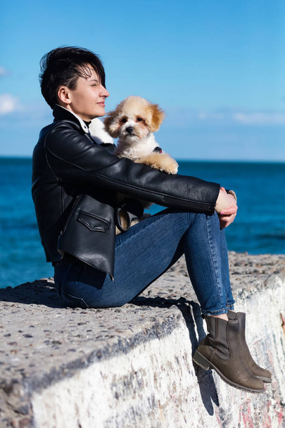Dreamy lady with stylish short hair and in fashionable skinny jeans, leather jacket. Sitting on the sea pier with the doggy. Small friend lying on the knees of the woman.Sunny day for the outdoor walk - Photo, Image