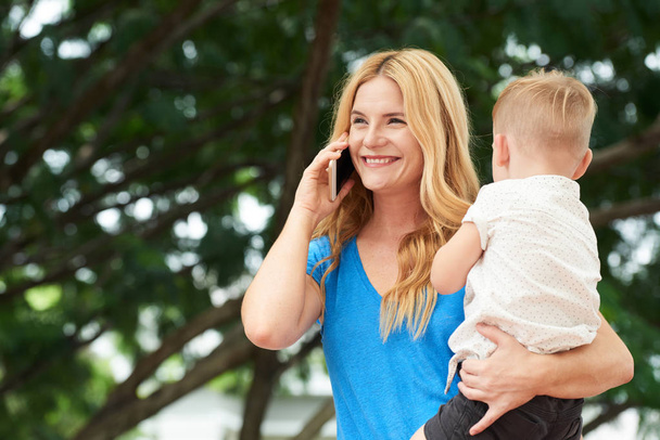 Mãe feliz carregando seu filho e falando ao telefone
 - Foto, Imagem