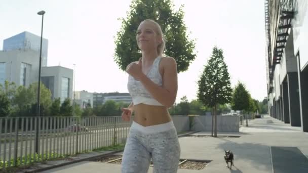 SLOW MOTION CLOSE UP LENSE FLARE Cheerful blonde girl jogging on city streets with dog on sunny morning. Smiling young woman working out before heading to work. Athletic female training for marathon - Video, Çekim