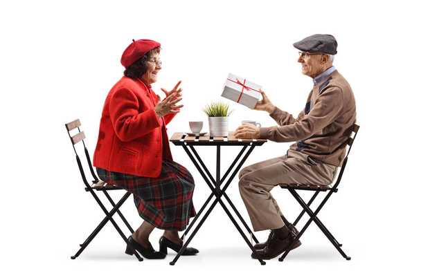 Full length profile shot of an elderly man sitting at a cafe and giving a present to an elderly woman isolated on white background - Foto, imagen