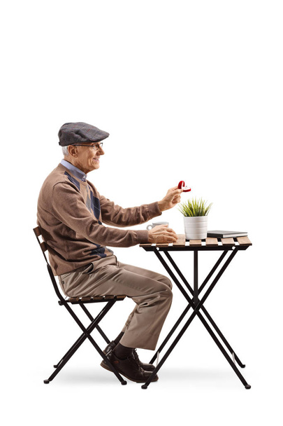 Full length profile shot of a senior man sitting at a cafe and holding a ring in a box isolated on white background - Photo, Image