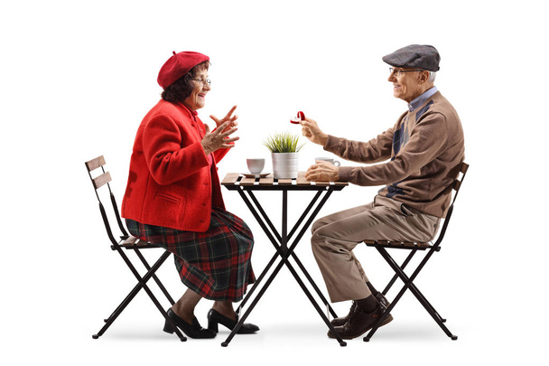 Volledige lengte profiel shot van een senior man zittend in een cafe en het geven van een ring aan een senior vrouw geïsoleerd op witte achtergrond - Foto, afbeelding