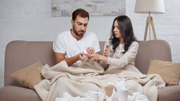brunette woman sneezing, bearded man giving tissue, coughing and woman taking glass of water and pills to sick man while sitting on sofa under blanket  - Footage, Video