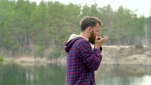 Young beard guy playing on the harmonica outdoors. Lake with forest at the background. - Кадры, видео