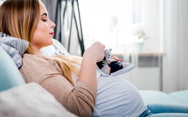Young pregnant woman with small baby shoes sitting on sofa at home, child expectation concept - Foto, Imagem