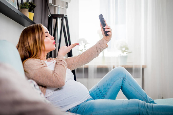 Mujer embarazada joven sentada en el sofá y tomando una foto selfie usando un teléfono inteligente
 - Foto, imagen
