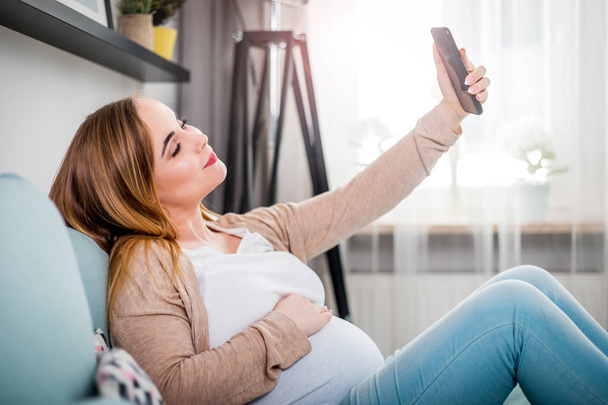 Young pregnant woman sitting on sofa and taking selfie photo using smartphone - Fotó, kép