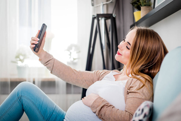 Jeune femme enceinte assise sur le canapé et prenant selfie photo à l'aide d'un smartphone
 - Photo, image