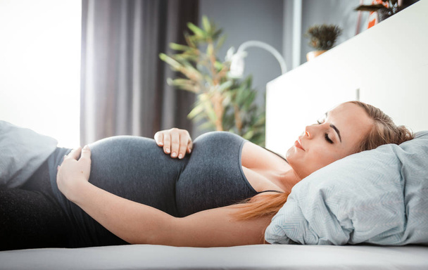Young pregnant woman sitting on bed and touching her belly, child expectation concept - Foto, Bild