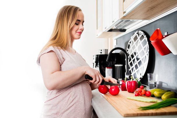 Pregnant woman preparing salad and cutting vegetables in kitchen - Фото, изображение
