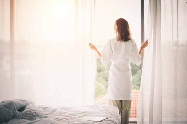 Bekijk terug vrouw opening gordijnen in een witte slaapkamer - Foto, afbeelding