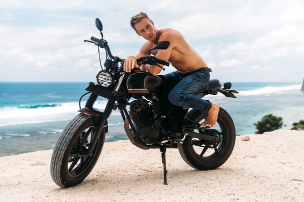 Sexy athletic man with perfect naked body sitting on motorbike, ocean waves and beautiful mountains on background - Foto, imagen