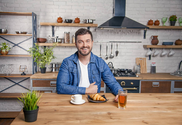 Nuori komea hymyilevä mies istuu keittiössä valmiina aamiaiselle. croissant, kahvia ja omenamehua
 - Valokuva, kuva