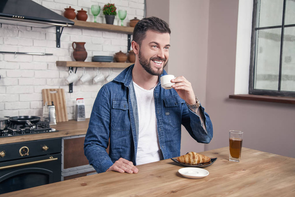 Genç yakışıklı gülümseyen adam kruvasan ve mutfakta bir elma suyu ile sabah kahve içiyor - Fotoğraf, Görsel
