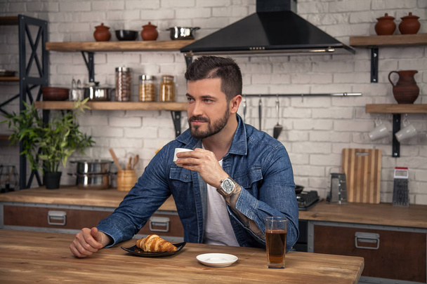 young handsome man is sitting at his kitchen in the morning having his coffee, croissant and an apple juice and thinking - Foto, imagen