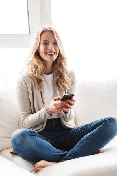 Beautiful blonde woman posing sitting indoors at home using mobile phone. - Fotó, kép