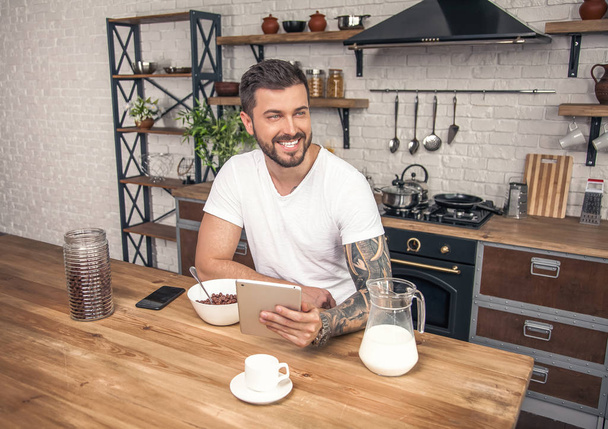 jovem bonito sorrindo homem está tendo seus cereais de pequeno-almoço com leite na cozinha e lendo notícias da manhã no tablet
 - Foto, Imagem