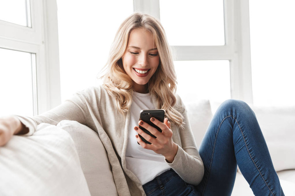 Beautiful blonde woman posing sitting indoors at home using mobile phone. - Photo, Image