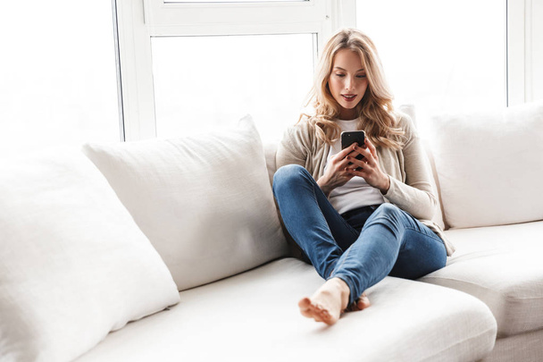 Beautiful blonde woman posing sitting indoors at home using mobile phone. - Foto, Imagem