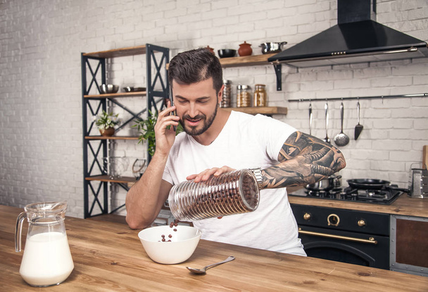 fiatal boldog jóképű mosolygó fickó készíti a reggelit a konyhában keverés gabona tejjel és miután telefon beszélgetés - Fotó, kép