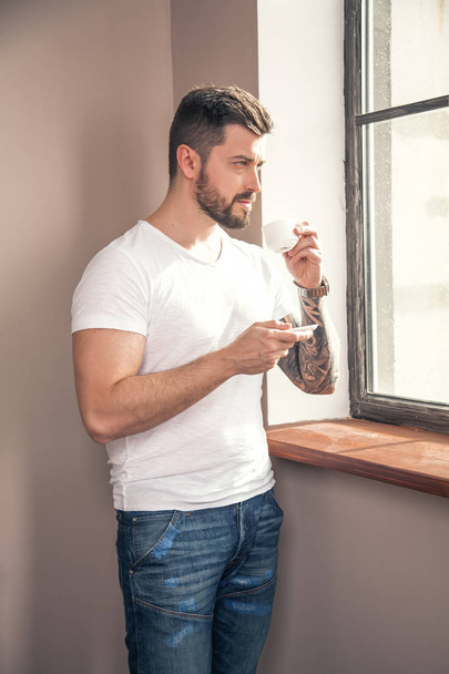 jonge knappe attente man is op zoek naar het raam en met zijn 's ochtends koffie - Foto, afbeelding