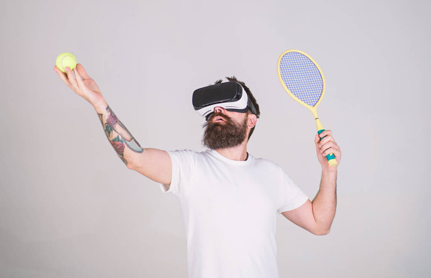 Handsome man in virtual reality headset playing tennis, grey background. Hipster use modern technology for sport games. Guy with VR glasses play tennis with racket and ball. Virtual tennis concept - Photo, Image