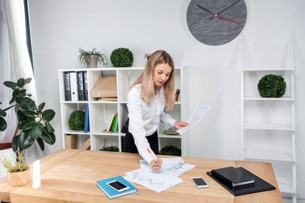 Theme business woman at work. Beautiful young caucasian woman business man working standing in the office near the table, checks edits financial charts on paper - Foto, Imagen