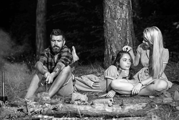 Bearded man roast sausage for women in vintage dresses. Hipster and girls relax at bonfire in forest. Boyfriend cook food on fire for girlfriends. Camping, cooking food and summer vacation - Foto, Imagem