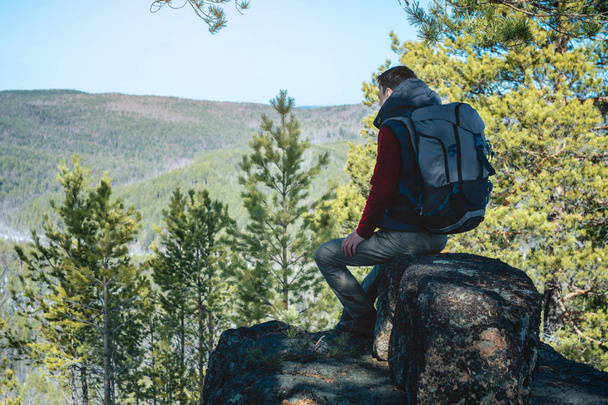 Ein Mann wandert mit einem großen Rucksack auf einem Felsen sitzend und blickt auf das weitläufige grüne Tal. Reisefreiheit - Foto, Bild