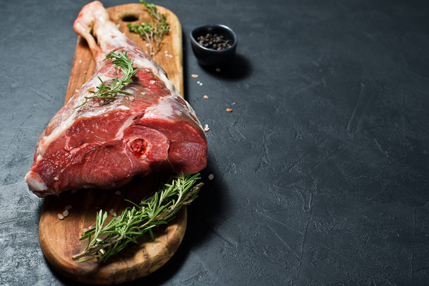 A raw leg of lamb on a wooden chopping Board. Rosemary, thyme, black pepper. Black background, side view, space for text - Valokuva, kuva