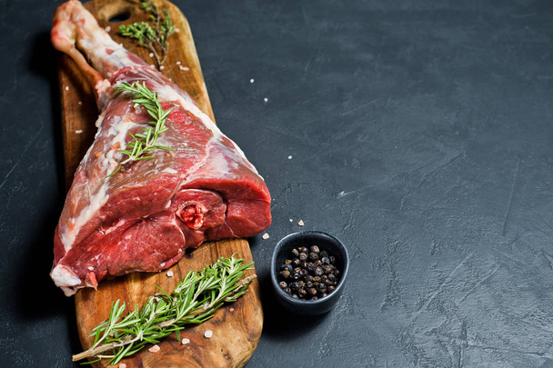 Raw leg of goat on a wooden cutting Board. Rosemary, thyme, black pepper. Black background, side view, space for text - 写真・画像