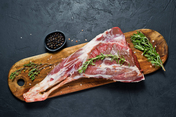 Raw leg of lamb on a wooden chopping Board. Rosemary, thyme, black pepper. Black background, top view - Photo, Image