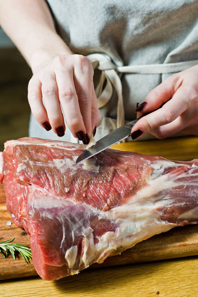 The chef cuts a raw leg of lamb on a wooden chopping Board. Rosemary, thyme, black pepper. Black background, top view, space for text - Photo, Image