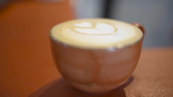 Κοντινό κομμάτι Κυπέλλου με καφέ και μοτίβο. Άρθρο. Ζεστό και αρωματικό καφέ κούπα με καρδιά μοτίβο της κρέμας είναι στο τραπέζι στο καφέ - Πλάνα, βίντεο