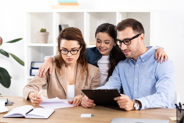 cheerful kid hugging mother and father in glasses looking at papers in office   - Foto, Imagen